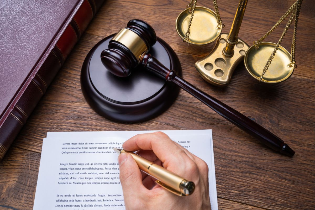 A person writing on a piece of paper next to a gavel and a balanced scale of justice on the table